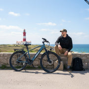 Man sitting next to a blue bicycle with a lighthouse and ocean view in the background