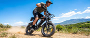 A man wearing a helmet and casual clothing rides a black electric fat-tire bicycle on a dusty dirt trail outdoors, with green hills and mountains under a blue sky in the background.