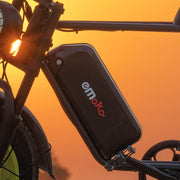 A close-up of a black emoka battery mounted on the frame of an electric bicycle, set against a warm orange sunset background.