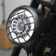 A close-up of a black industrial spotlight with a protective cage over a bright white LED light, mounted on a black metal frame.