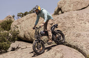 Person riding an electric bike on rocky terrain with large boulders and a clear sky.