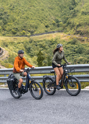 A man in an orange hoodie and a woman in an olive green top ride electric bicycles side by side on a paved road with a lush green mountain backdrop.