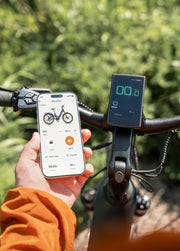 A person holds a smartphone showing a bike app screen in front of a bicycle handlebar with a mounted display. The background is a blurred outdoor green setting.