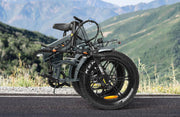 A folded electric bicycle with thick knobby tires and a dark gray frame, parked on an asphalt road with a mountain landscape in the background.