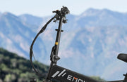 Close-up of a black electric bicycle frame and handlebar area with visible cable routing, set against a blurred mountain landscape background. The frame shows partial branding text 'EMO'.