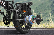 Close-up rear view of an electric bicycle on a road with a blurred green hillside background. The bike has a black frame and fat tire, with a visible exposed rear motor showing blue, green, and orange internal components.