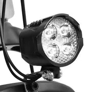 A close-up of a black bicycle headlight with four clear LED lights, mounted on a bike handlebar with wiring visible against a white background.