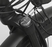 Close-up of a black electric bicycle frame and handlebar area with a silver throttle or control knob, attached cables, and a metal mounting bracket against a light gray background.
