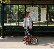 A woman stands in a park beside a red folding bicycle under a shelter, with trees, benches, and informational signs in the background.