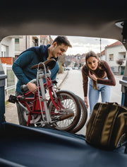 A man and a woman loading a red folding bicycle into the trunk of a car, with a brown bag in the foreground and a suburban street in the background.