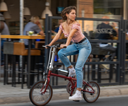 A woman rides a compact red folding bicycle on a city street in front of an outdoor cafe, wearing a pink ruffled top, blue jeans, and white sneakers.